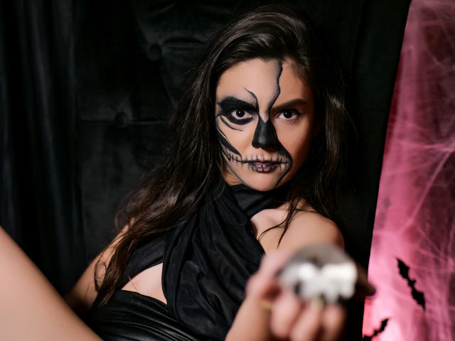 AshlyChloe avec un maquillage foncé, de la peinture noire sur le visage, une robe noire, tenant un papillon blanc, des cheveux bruns ondulés, un fond noir et rose, regardant la caméra, expression sérieuse..