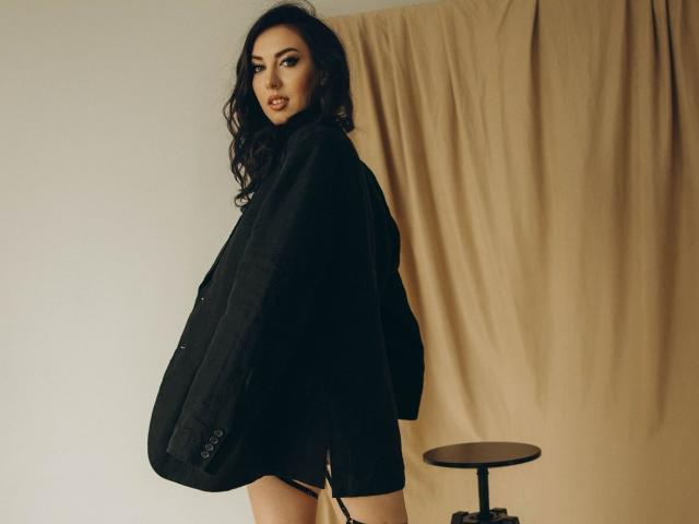 KaleyReed with wavy dark hair, wearing a black jacket, looking over her shoulder, standing next to a small round stool, tan curtain in the background.
