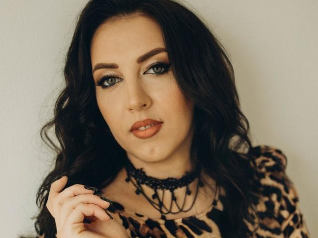 KaleyReed has dark wavy hair, blue eyes, and a leopard print shirt. She's wearing black lace chokers and has a slightly pouty mouth with orange lipstick.
