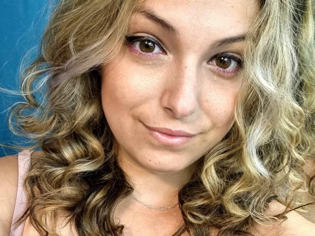 MissAnalis heeft golvend blond haar, bruine ogen met roze eyeliner, en een subtiele glimlach. Ze draagt een roze top en een dunne ketting. Haar borsten zijn niet zichtbaar..