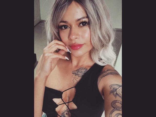 DariaDawson, con capelli grigi sfumati, una maglia nera con lacci, rossetto rosso, tatuaggi sul petto e sul braccio, si tiene il mento, sorride, mostrando il seno..