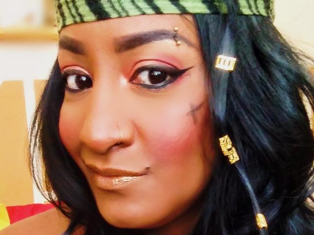 Nahirr's face with dark brown skin, black hair, green striped headband, gold earrings, red eyeshadow, black winged eyeliner, nose piercing, lip piercing, and a gold lip color.