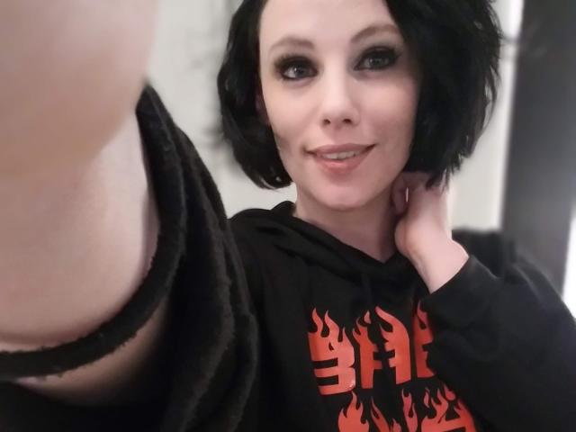 KylieRyder, con i capelli neri corti, indossa una felpa nera con la scritta "Bleed", sorride alla fotocamera, si tiene il collo, mostrando parzialmente i suoi seni grandi e scoperti sul lato sinistro..