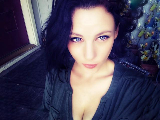 KylieRyder, con cabello negro ondulado, ojos azules y una camisa negra ligeramente abierta que muestra su escote, mirando hacia la cámara con una sonrisa sutil y sensual..