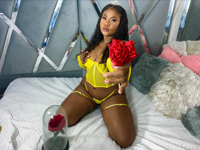 Sara Lourens, com pele escura, vestindo lingerie amarela, segurando rosas vermelhas, sentada em uma cama com lençóis brancos, um copo com uma rosa e almofadas rosa ao fundo..