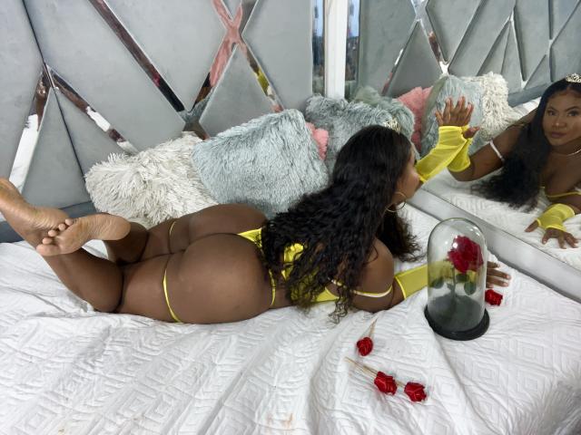SaraLourens ligt op een bed, haar gezicht in de spiegel, gekleed in gele lingerie, handschoenen en een string, met krullend zwart haar, en haar blote billen, met haar voeten omhoog, rode rozen in een glazen koepel..