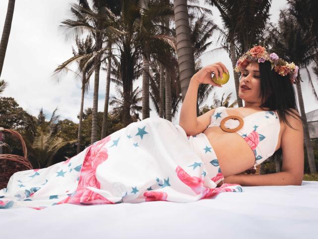 WendySchulz lying on a white blanket, wearing a star-patterned top and pink skirt, holding a yellow apple, with a flower crown, showing her tits, in a tropical setting.