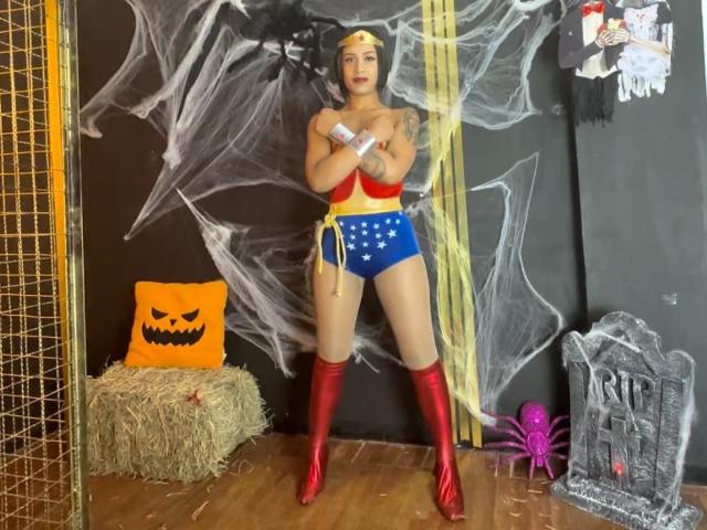KendallQuinn in Wonder Woman cosplay, standing with arms crossed, wearing red boots, blue star-spangled shorts, yellow top, red bustier, yellow headband, against black wall with spider webs.