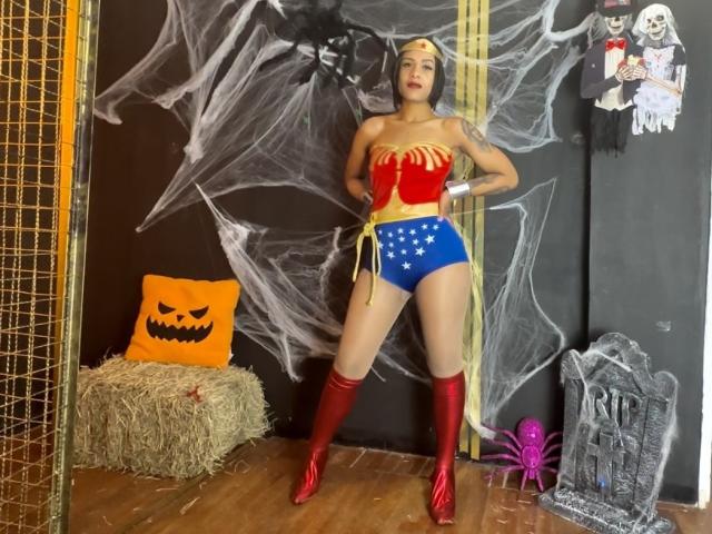 KendallQuinn in Wonder Woman kostuum met rode laarzen, blauwe shorts met sterrenpatroon, een rood en geel top en een gele riem, staand met handen op de heupen, waarbij haar billen en borsten zichtbaar zijn..