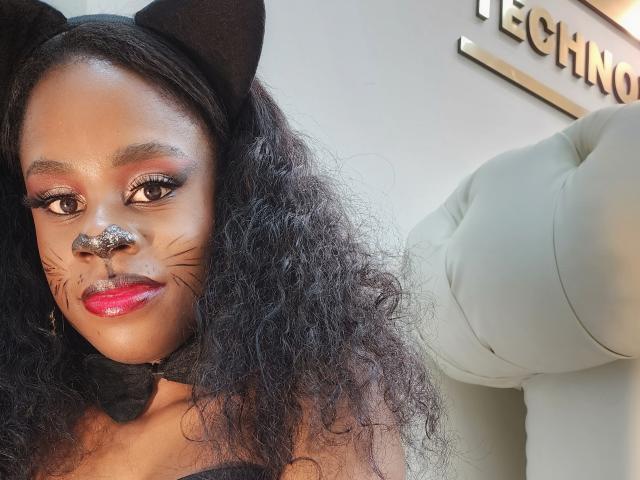 MiaHopson with curly black hair, black cat ears, white nose paint, pink lipstick, and black bow, wearing a cat face mask, smiling, showing her ass in a white chair.