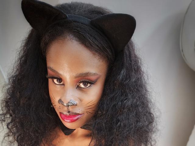 MiaHopson with dark curly hair, black cat ears, glitter nose, fake whiskers, red lipstick, and smoky eye makeup, looking up with a cute, playful expression.
