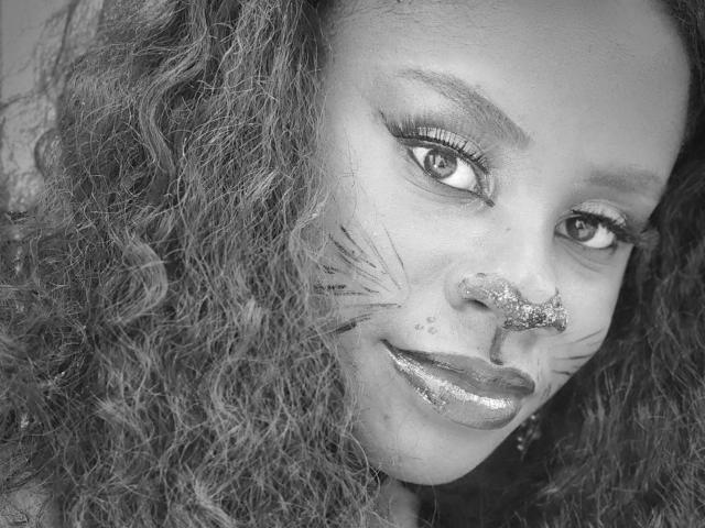 MiaHopson's face with glitter on her nose and "whisker" lines on her cheeks, curly hair, glossy lips, and subtle makeup, in black-and-white, staring confidently at the camera.