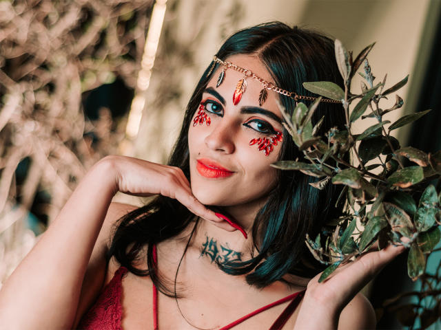CoriPaige, con i capelli scuri, il rossetto rosso e il trucco rosso sul viso, indossa una corona dorata e una camicetta di pizzo rossa, tiene in mano un ramo di foglie e mostra un tatuaggio sul collo..