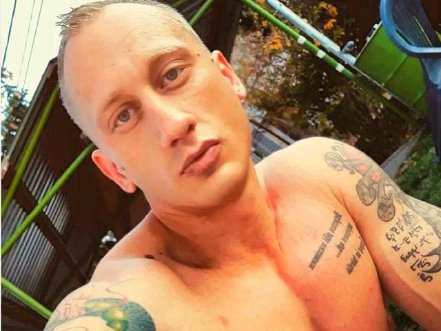 TonyLove69, ohne Hemd und mit Tattoos, hat einen rasierten Kopf, helle Haut und blaue Augen. Er blickt mit leicht zusammengekniffenen Lippen in die Kamera. Seine Tattoos zeigen ein Fahrrad und Text..