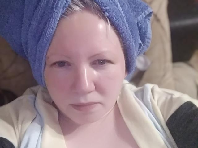Agnnessa heeft een blauwe handdoek op haar hoofd, een glanzend gezicht en een neutrale uitdrukking. Ze draagt een beige en zwarte top. Haar huid is bleek..