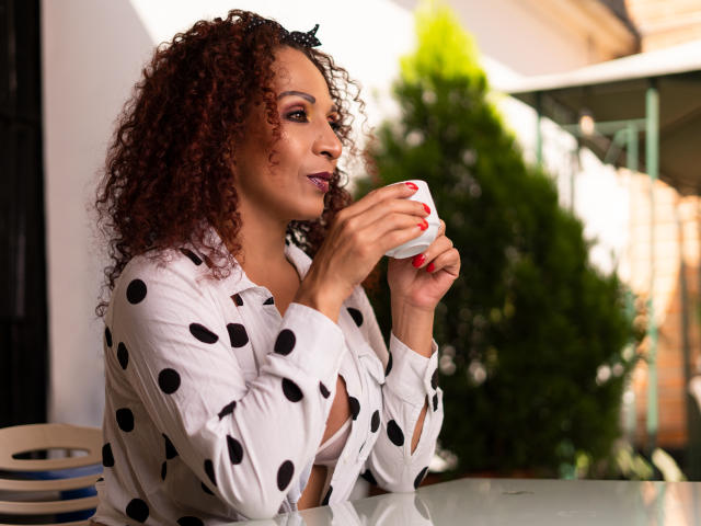 KhazandraX with curly brown hair, wearing a white polka dot shirt, sips from a white cup with red nails, smiling slightly, sitting at a glass table.