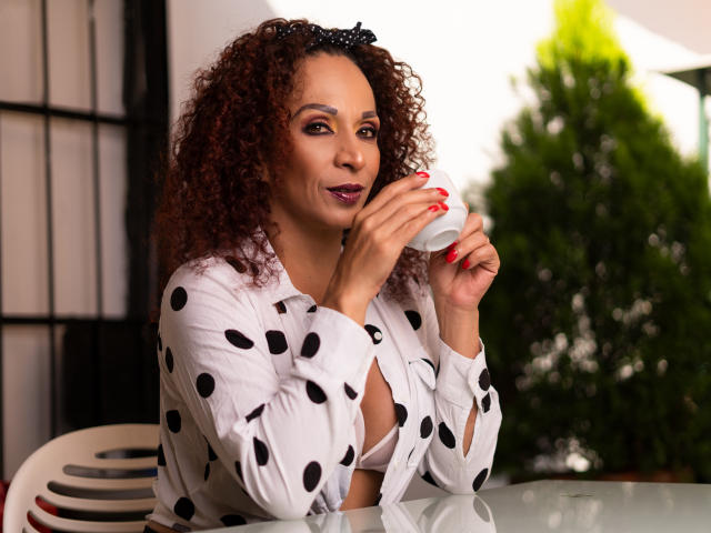 KhazandraX with curly red hair, polka dot shirt, sipping from a cup, showing red nails, slightly open shirt revealing white bra, smiling subtly, green plant in background.