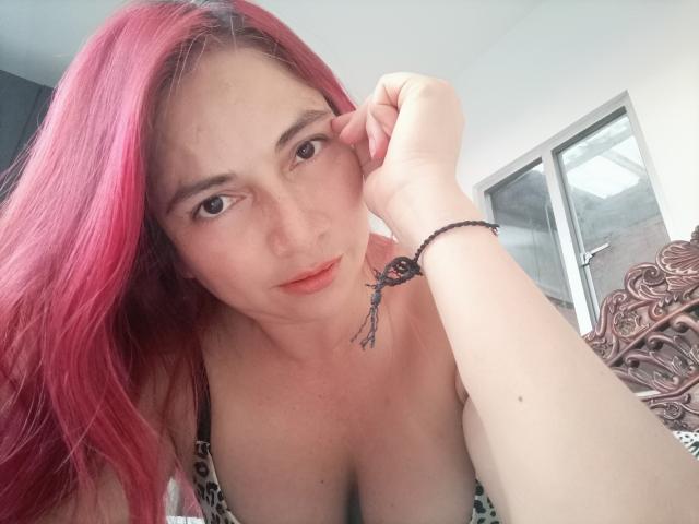 AlessiaRed with pink hair, brown eyes, and red lips, looking at the camera, showing her tits in a leopard print bra, wearing a black bracelet with a feather.