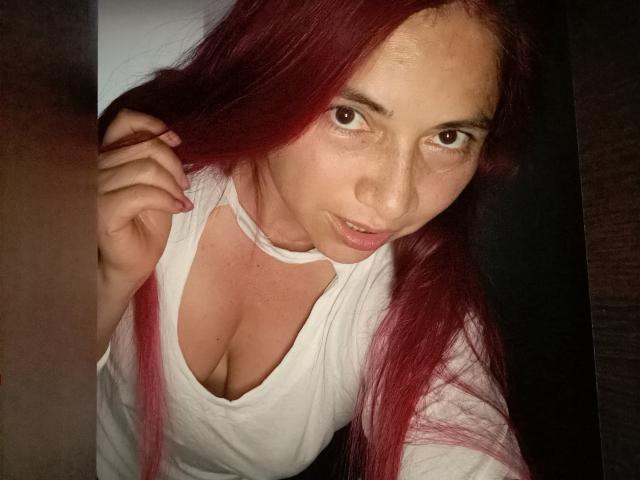 AlessiaRed with red hair, white shirt, and cleavage, holding her hair, looking at the camera, with a slightly open mouth and a cute expression.