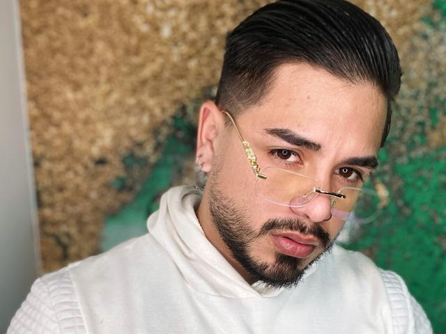 VisiI tem barba, cabelo preto curto e um piercing no nariz. Ele usa óculos transparentes com armação dourada e um suéter branco texturizado de gola alta. O rosto de VisiI demonstra uma expressão séria..