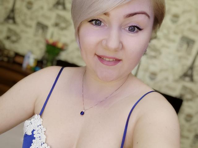 BlondPearlX con i capelli biondi corti, occhi azzurri e un sorriso, indossa un reggiseno di pizzo blu, una collana con una pietra blu e mostra il seno in un selfie..