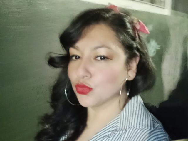 SakotoDika with red lipstick, hoop earrings, and a pink bow in black hair, wearing a blue-striped shirt, pouting slightly.