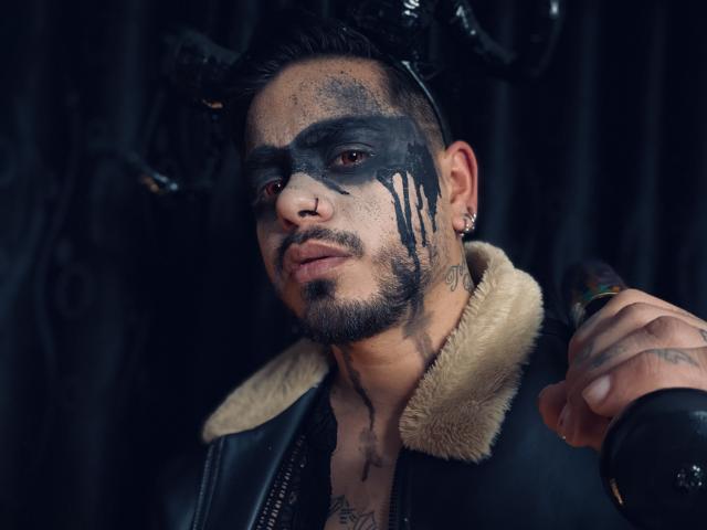 VisiI tem maquiagem preta no rosto, uma coroa de chifres e um casaco de couro com gola de pele, segurando um morcego. Ele tem tatuagens, um piercing no nariz e barba. VisiI parece intenso..