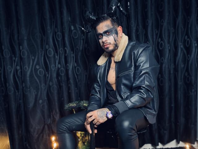 VisiI con chaqueta de cuero negro, cuello de piel, maquillaje facial negro, tocado de cuernos negros, pantalones negros, manos tatuadas, sentado, fondo de cortina oscura, textura de cuero, expresión seria, reloj de pulsera..