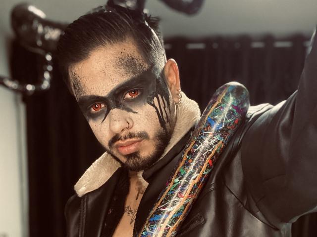 VisiI a du maquillage noir sur le visage, des yeux rouges, et une veste en cuir noir. Il porte une batte de baseball colorée, avec une peinture écaillée, en bandoulière. Les cheveux de VisiI sont peignés en arrière..