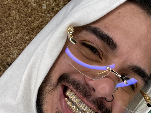 Il viso di VisiI, coperto da un velo bianco, indossa occhiali con filtro per la luce blu, ha un sorriso con denti dorati e un piercing al naso, appare felice con un leggero sorriso..