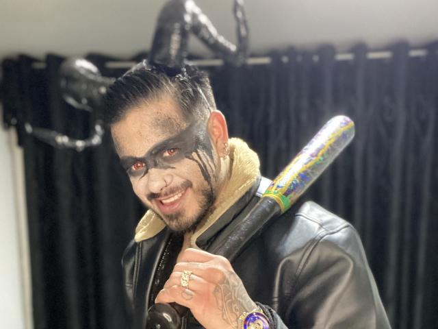 VisiI, avec du maquillage noir sur le visage, une veste en cuir et un bat, le tenant sur son épaule, souriant, a des tatouages sur sa main droite et porte une bague. Un rideau sombre est visible en arrière-plan..