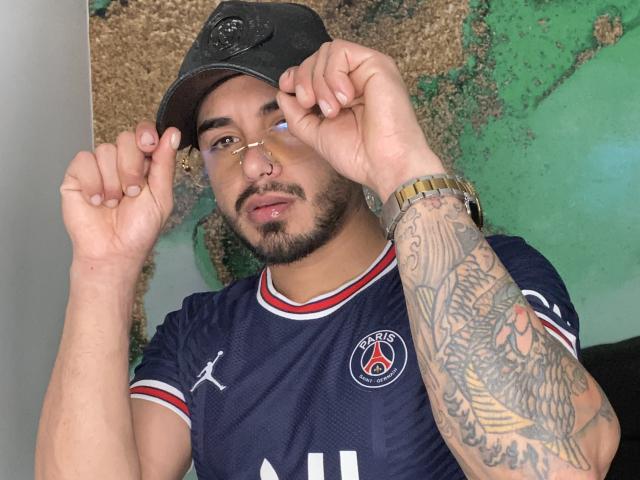 VisiI, com tatuagens no braço direito, usando um boné preto, óculos, piercing no nariz e uma camisa de futebol Puma, ajustando o boné. VisiI tem barba e um relógio de ouro..