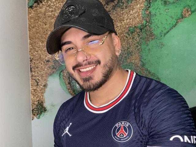 Um homem com óculos, piercing no nariz e barba, vestindo um boné preto e uma camisa de futebol PESCE azul marinho, sorrindo para a câmera. A camisa dele tem uma borda vermelha, branca e azul..