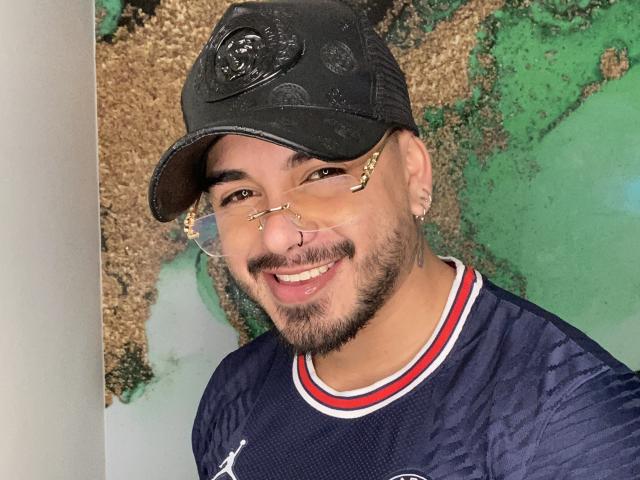 VisiI lleva una gorra de béisbol negra, gafas con montura dorada, un piercing en la nariz y un bigote, y viste una camiseta azul marino con detalles en rojo y blanco, sonriendo a la cámara..