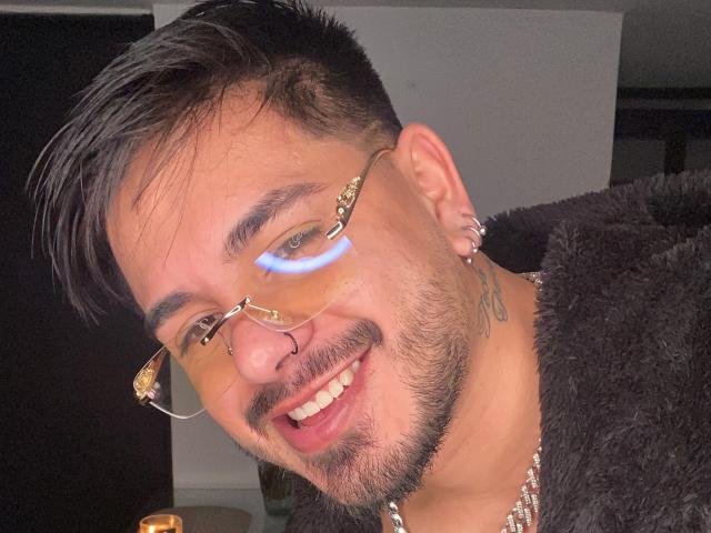 A VisiI tem um sorriso, usa um piercing no nariz, óculos e tem uma tatuagem, vestindo uma jaqueta preta de pelos e um colar de corrente, com um corte de cabelo preto curto e brincos..