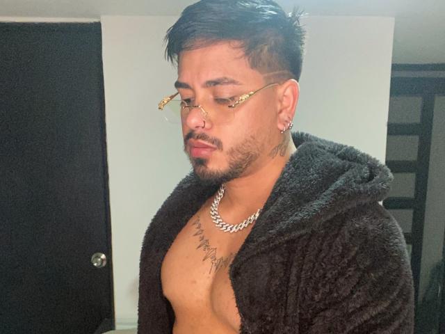 VisiI, com cabelo preto curto, óculos e piercing no nariz, vestindo um robe preto felpudo, com o peito tatuado à mostra, e uma corrente de prata, em pé em um quarto com pouca luz..