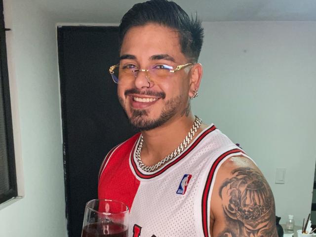 VisiI hat dunkles Haar, eine Brille und einen Bart, trägt ein rotes und weißes Basketballtrikot mit einem Tattoo auf seinem rechten Arm und hält ein Glas mit einem roten Getränk..