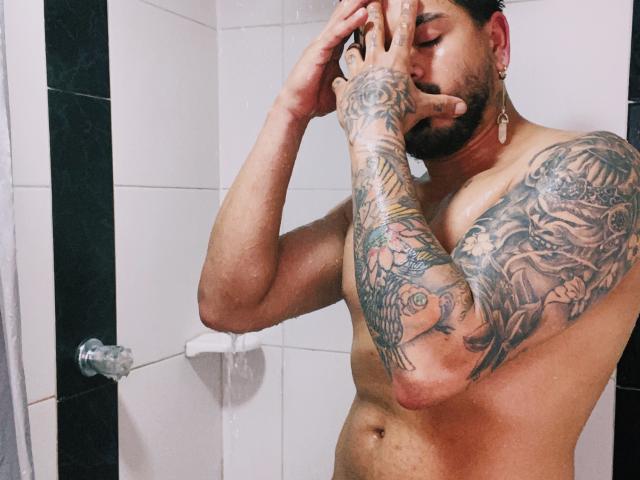 VisiI, ohne Hemd und mit Tattoos auf Arm und Brust, steht unter der Dusche, wobei er sein Gesicht mit einer Hand bedeckt. Wasser tropft von seinen kurzen, dunklen Haaren und seinem Bart..