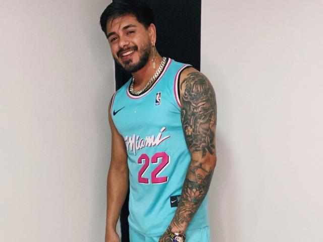 VisiI, een man met tatoeages en een baard, draagt een blauw basketbalshirt van de Milwaukee Bucks (#22), glimlacht en staat tegen een witte muur, terwijl hij een gouden ketting draagt..
