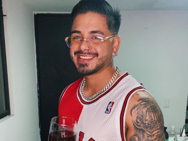 Uma pessoa do sexo feminino, com cabelo curto e escuro, óculos, piercing no nariz e barba, sorrindo, vestindo uma camisa de basquete vermelha e branca, segurando um copo com líquido vermelho, com tatuagem no braço direito..