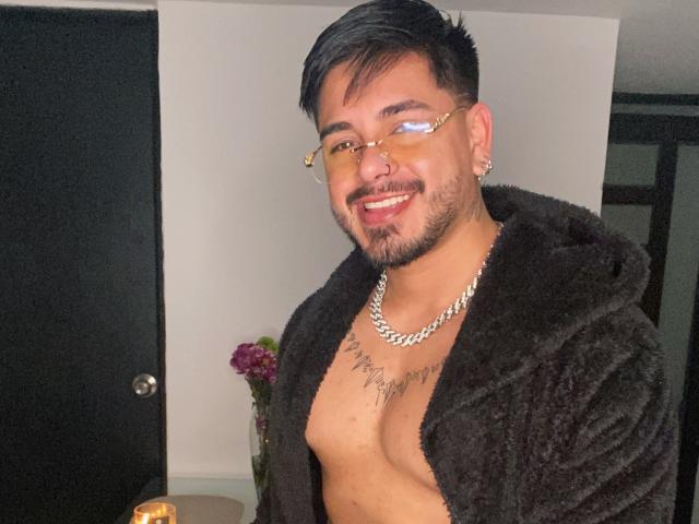 VisiI tem uma pelagem escura, óculos dourados e um colar de corrente, sorrindo com barba, tatuagem no peito e peito exposto, segurando uma bebida..