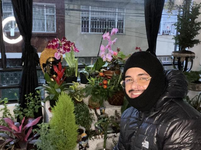 VisiI con gli occhiali e una balaclava nera, indossa un giubbotto nero imbottito, sorride e sta in piedi tra fiori colorati, tra cui orchidee rosa e gigli rossi. VisiI ha i baffi e indossa un cartellino con il nome..