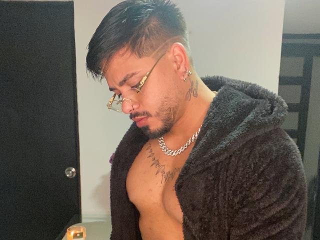 VisiI, sin camisa y con una sudadera negra de pelo, tiene tatuajes en el pecho, una cadena de oro y gafas. El cabello oscuro de VisiI está peinado hacia arriba, y VisiI está mirando hacia abajo con una expresión seria..