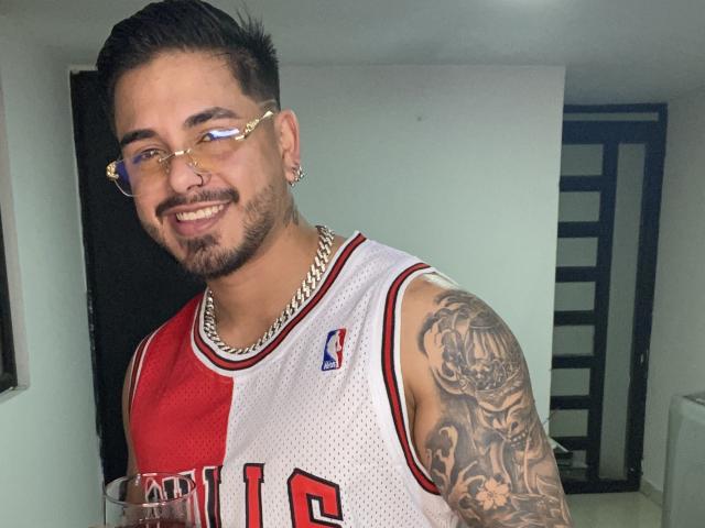 VisiI, con capelli scuri e corti, occhiali e un piercing al naso, indossa una maglia da basket bianca e rossa, mostrando un braccio tatuato, sorride e tiene in mano un drink..