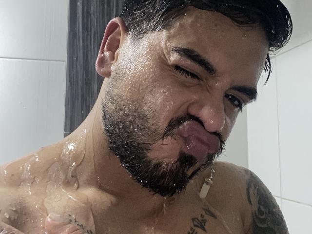 VisiI hat ein feuchtes, bartbewachsenes Gesicht mit kurzen, dunklen Haaren, steckt seine Zunge heraus und Wasser läuft ihm über die Brust und die Tattoos. Seine Augen sind leicht zusammengekniffen und halb geschlossen..