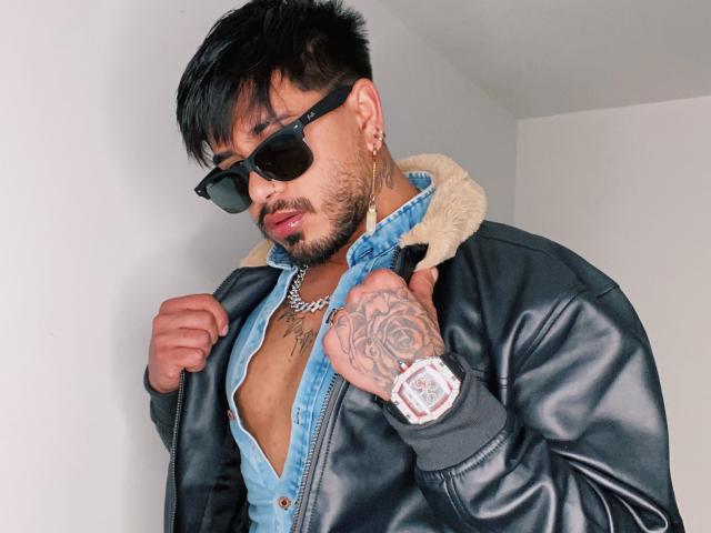 Eine Person mit dunklem Haar, Sonnenbrille und Bart, die eine schwarze Lederjacke über einem offenen, blauen Hemd trägt, wobei Tätowierungen auf Brust und Hand sichtbar sind, und eine silberne Uhr..