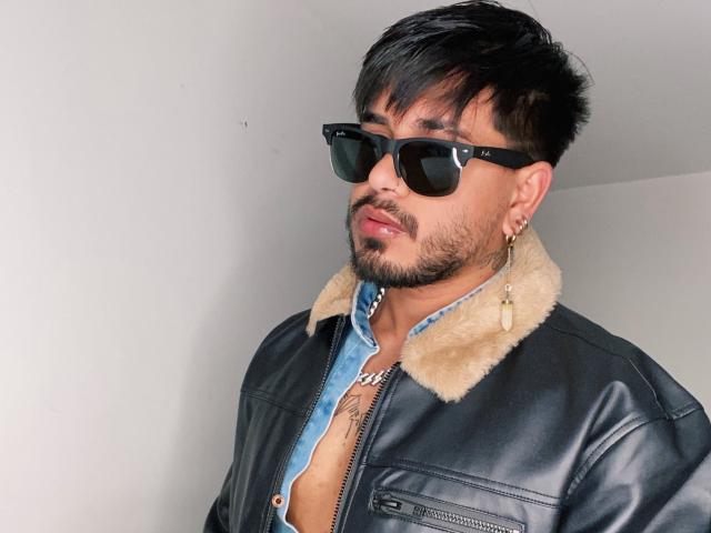 VisiI con occhiali da sole neri, una giacca di pelle nera con un colletto in pelliccia color cuoio, e una camicia blu, che mostra un tatuaggio sul petto, e con un orecchino d'oro..