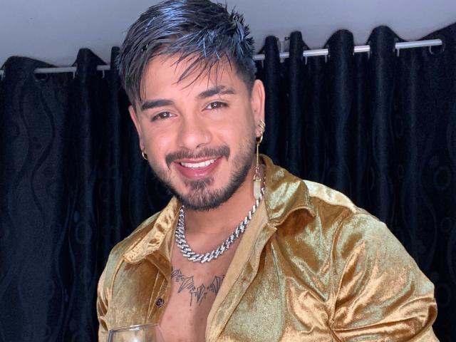 VisiI has a shiny gold shirt, a silver chain, and a chest tattoo, smiling with short, spiked black hair and a beard. He's holding a clear glass.