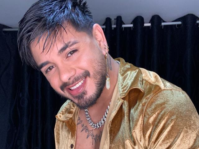 VisiI tem um sorriso, cabelo preto curto com mechas azuis, barba e está vestindo uma camisa dourada brilhante com um colar de prata. Ele tem tatuagens no pescoço..