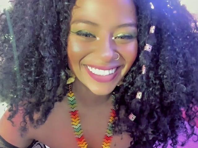 NashaJonny with dark curly hair, green eyeshadow, pink lipstick, nose ring, smiling, colorful necklace, showing her tits, pink top, glowing under soft light.